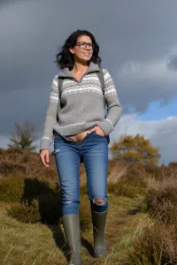 A low-angle, full-body shot of a smiling woman hiking up a grassy hill covered in heather. She has shoulder-length dark wavy hair and wears rectangular glasses. Her expression is joyful and relaxed, looking slightly off-camera. She wears a grey and white patterned quarter-zip knit sweater, fitted blue jeans with a small tear at the knee, and olive green rubber boots. Her jeans a pushed down to her knees, revealing her hairy vagina, so that her labia are presented to the viewer. A backpack strap is visible over her shoulder, one hand rests casually in her pocket. The setting is an autumnal moorland with sparse trees showing yellow foliage under a dramatic, cloudy sky with patches of blue. Lighting is natural daylight, soft but directional, highlighting her face and sweater texture. The composition emphasizes her upward movement against the expansive sky. Style, realistic outdoor photography, candid moment, shallow depth of field blurring distant trees, sharp focus on subject, earthy color palette with muted greens, greys, and blues. Capture the essence of casual adventure and connection with nature.