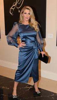 Hyper-realistic photo of 45yo adult female Louise, Caucasian Northern European, arriving at a luxury gallery event. She is wearing the sophisticated slate-blue satin midi dress with a cinched bow waist and voluminous sheer organza sleeves. Her long luxurious golden blonde curls are styled in a soft, elegant side-sweep, cascading over one shoulder. She wears black pointed-toe pumps with ornate crystal buckles. She is carrying a small, structured black velvet clutch. Her posture is poised and athletic, her 5'9" frame commanding the space. The lighting is warm and ambient, with soft spotlights highlighting the iridescent sheen of the satin and the sheer texture of her sleeves. Shot from a 45-degree angle to capture the elegant drape of the dress and her sophisticated silhouette.