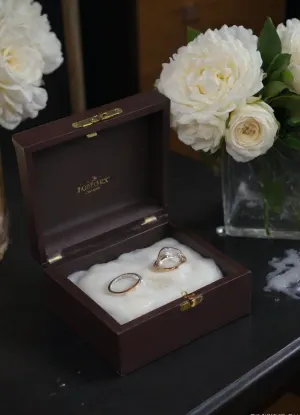 A surreal close-up of two wedding rings inside an open, elegant ring box placed on a black velvet table. Beside the box lies a delicate white bridal bouquet. Thick, glossy white slime with creamy undertones is slowly poured over the box and flowers, dripping down the sides and partially covering the rings. The lighting is soft and cinematic, enhancing the shine of the rings, the texture of the slime, and the dark contrast of the background. The scene feels poetic, artistic, and symbolically messy—blending elegance with surreal distortion.