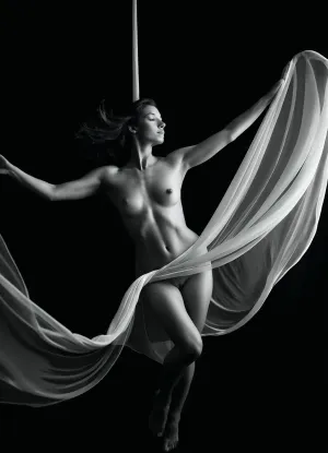 A black and white fine-art nude photography-style image of a woman suspended within flowing, sheer silk fabric that lifts and holds her in midair. The delicate, translucent material wraps around her body, contouring her natural form while creating