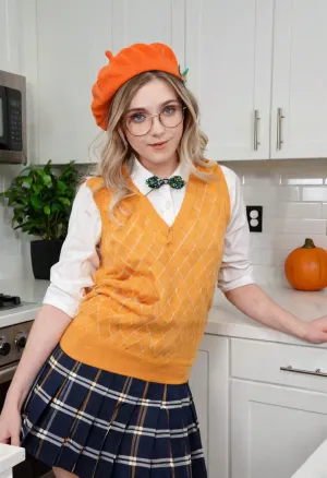 A young Caucasian woman, appearing to be in her early twenties, stands in a modern kitchen. She wears an orange beret with a small green leaf accent, round eyeglasses with a light brown frame, a white collared shirt beneath a yellow argyle sweater vest with brown ribbons and a pearl brooch. Completing the outfit is a navy blue pleated skirt. Her long, wavy blonde hair cascades over her shoulders. She gazes directly at the viewer with a friendly, slightly surprised expression. Her right hand rests on a white countertop. The kitchen features white cabinets with black handles, a stainless-steel refrigerator, a backsplash with white tile, wooden cutting boards, and a small plant. The lighting is bright and natural, giving the image a clean, inviting aesthetic. The scene's composition is balanced, using a medium shot to focus on the woman and her attire. The overall mood conveys a sense of youthful energy and modern domesticity. The style is reminiscent of a lifestyle photograph, with high-definition and soft lighting, creating an attractive image.