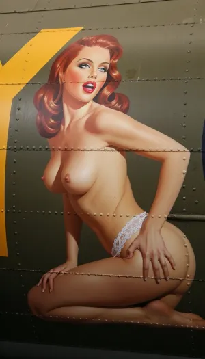 A vibrant and authentic vintage nose art painting stenciled onto the weathered olive-drab aluminum fuselage of a World War II B-17 bomber. The artwork depicts a stunning 20-year-old redhead pin-up girl with a youthful face and exquisite features, painted in a lush oil style with bold brushstrokes. She is posed kneeling in a side profile view, completely topless, with her bare breasts visible from the side. She wears only delicate retro lace lingerie bottoms. She has a playful, knowing expression with classic red lips and perfectly styled retro wavy hair. The painting is framed by metal rivets, panel lines. Vibrant saturated colors, perfect vintage pin-up proportions, bold outlines suitable for military aircraft insignia, and high-contrast lighting that makes the artwork pop against the matte metallic background.