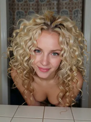 A young woman with voluminous blonde curly hair gazes at the camera with a playful smile, framed by a simple interior background.

The composition is a close-up portrait, focusing primarily on the woman's head, shoulders and chest. She is positioned centrally, The framing is tight, emphasizing the texture and volume of her hair, which cascades around her like a halo. There is no discernible action or interaction, as the image captures a static pose. The background elements are blurred and indistinct, serving to isolate the subject and draw all attention to her.

The key subject is a young woman with very fair pale skin and striking, voluminous blonde hair styled in tight, natural curls. Her eyes, light in color, mischievous expression. playful or thoughtful mood. She is topless, with small breasts. Her makeup is minimal, highlighting her natural features.



The setting is an indoor environment, suggested by the presence of what looks like a patterned curtain or backdrop behind her, and a tiled surface in the foreground that resembles a countertop or table. The overall atmosphere is relaxed and informal. The lighting is not dramatic, it appears to be ambient room lighting, which creates a soft, even illumination. This subdued lighting contributes to the intimate and personal feel of the portrait. The mood is one of gentle curiosity or lightheartedness, conveyed through her expression and the framing of her gaze.