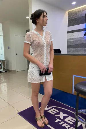 A woman stands in profile at a reception desk in a modern, brightly lit interior, captured in a medium full-body shot. She has short, dark brown hair styled in a casual updo with a black hair clip, and her face shows a neutral expression as she looks toward the right. She wears a white, short-sleeved transparent dress with a subtle black polka dot pattern, featuring a button-front detail. Her small breasts, with their erect nipples, are visible through the sheer dress. In her hands, she holds a small, dark clutch or wallet. Her footwear consists of metallic gold or bronze-colored sandals. The setting is a clean, contemporary lobby with a tiled floor, a wooden reception counter with blue geometric accents, and a textured gray and white wall. A purple mat with "NEXEN TIRE" branding lies on the floor. The lighting is even and bright, typical of indoor commercial spaces, creating soft shadows. The overall style is realistic and candid, resembling a natural photograph with a clear focus on the subject and her immediate environment.