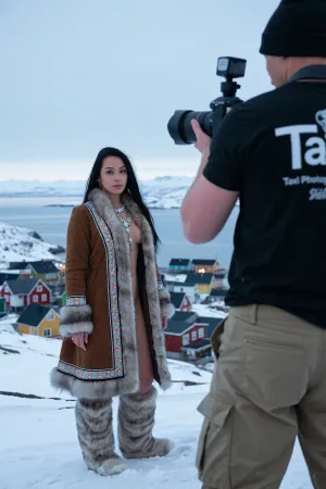 A photorealistic cinematic fashion photoshoot on a snow-covered hill in Greenland, bathed in soft pale daylight, the vast arctic landscape stretching toward a small village with brightly painted wooden houses in vivid colors, nestled along a fjord, the sky expansive and crisp, film noir atmosphere with a cool blue tone, a subtle mist in the distance softening the mountains, the colorful city Nuuk below gently illuminated by the crisp light, the photographer visible in the foreground from an over-shoulder perspective, waist-high, holding a professional camera, he wears khaki trousers and a black T-shirt with a white DSLR logo and the text “Taxi Photography, ” his shoulders, camera, and head slightly blurred, slightly blurred to create cinematic depth of field, The main focus is a tall Inuit female model, 22 years old, with classic Inuit features, a serene, confident expression, long black hair flowing in the cold wind, wearing traditional Inuit attire with intricate beadwork and fur trim, a brown fur-lined parka open to reveal her graceful form beneath, she poses with quiet strength, standing tall on the snow, gazing over her shoulder toward the camera with a subtle, poised smile, every detail of her skin, the fur, and beadwork rendered in hyper-realistic clarity, subtle shimmer of frost, masterpiece composition, ultra-realistic lighting and textures, luxurious fashion noir aesthetic, maximum realism, emotionally cinematic visual storytelling