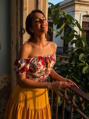A striking image of a beautiful Cuban woman standing on a balcony in Old Havana, bathed in the warm glow of a sunset. She has her eyes closed, a serene smile on her face, savouring the golden rays of the sun on her sun kissed skin. Her attire consists of a vibrant Camisa de Campesina with intricate floral patterns, worn off the shoulders, and a flowing yellow Pollera skirt. The composition should include lush green foliage and old weathered walls of the colonial architecture in the background, highlighted by the rich, warm colours of the sunset. The atmosphere is joyful and tranquil. Aim for high resolution, to capture every detail vividly.
