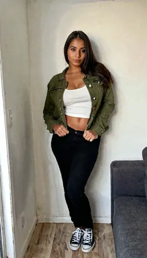 Ultra-realistic smartphone photograph of a 25-year-old adult Hispanic woman with a naturally slim-curvy feminine figure, warm olive skin, long dark brown hair and expressive brown eyes. She is wearing a denim jacket over a white top with black trousers and casual sneakers. She is leaning casually against a wall in a minimalist living room, looking directly at the camera with a relaxed expression. Natural indoor daylight, handheld smartphone camera, realistic exposure, authentic skin texture, detailed denim fabric, natural shadows, true-to-life colors, candid photography, highly photorealistic.