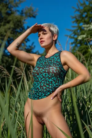 A medium-full outdoor shot of a woman standing in tall marsh grass, shielding her eyes from the sun with her left hand while her right hand rests on her hip. She has short, platinum blonde hair with dark roots and a focused, squinting expression as she gazes into the distance. She wears a sleeveless, patterned green-and-black top, but is naked underneath, (((displaying her very hairy vagina))), . A tattoo is visible on her upper left arm. The scene is bathed in bright, direct sunlight under a clear blue sky, casting sharp shadows. Lush green reeds and trees form a dense natural background. Photorealistic style, candid travel photography, high detail, vibrant colors, shallow depth of field emphasizing the foreground subject.