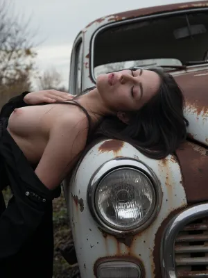a young woman leaning against the bonnet of an old rusted abandoned car, head tilted back, eyes half closed, expression distant and languid, long dark hair loose and windswept, breasts exposed, black clothing dissolving into the dark foreground, the car heavily rusted, pale paint peeling, round vintage headlight prominent in the frame, the car filling the background behind her, overcast flat grey sky, bare autumn trees dissolving into soft bokeh, cool and muted tones, rust and pale grey palette, analogue film, heavy grain, scratches and film imperfections, Kodak Portra 400 underexposed, cool and melancholic, shallow depth of field, the woman sharp against the soft dissolved background, quietly devastated, the rusted car as metaphor