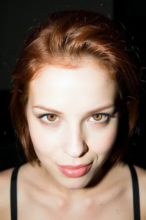 A striking portrait of a mischievous woman. A direct, hard camera-flash illuminates her face from close range, creating an overexposed, high-key skin tone against a pitch-black, dark void of a room. This creates strong contrast and a gritty, grainy film texture. Warm golden-brown eyes with sharp limbal rings and reflected catch-lights. Her short red hair is thick and voluminous styled in a chic swept-back look. Black bra straps contrast sharply with her pale skin. All background detail is swallowed by the darkness.