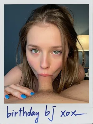 Mans POV. A close-up, intimate portrait of a young woman with fair skin and green eyes, her face positioned on a man's torso, her mouth just above his penis. She has long messy Beliage hair that frame her face. Her expression is direct and engaging, with wide eyes and slightly parted lips. She wears bright blue nail polish. The composition is tight, focusing on her face and upper torso as she leans forward, cum shooting into her mouth. The lighting is soft and warm, likely from a nearby lamp visible in the background, casting a gentle glow on the scene. The background is a simple, dark blue wall, keeping the focus on the subjects. The overall style is realistic and intimate, with a shallow depth of field that blurs the background slightly., ((Polaroid Photo handwritten signed with sharpie "birthday bj xox"))