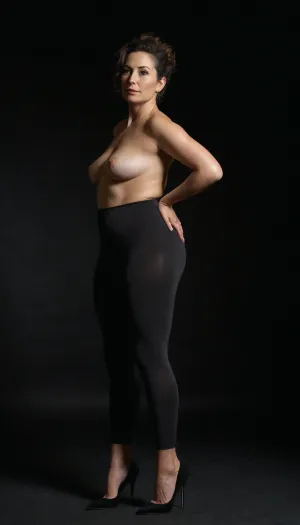 A full-length high-fashion editorial photograph of a poised 40-year-old woman with a dramatic, exaggerated pear-shaped silhouette. She has a slender, flat saggy chest that contrasts sharply with wide, opulent, curvaceous hips and a full, rounded posterior. She is dressed in understated, casual everyday attire—a simple fitted cotton tee and tight-fitting leggings—that clings to her form. She stands poised in black, needle-thin stiletto high heels that dramatically arch her feet. The lighting is cinematic rim lighting, carving her lower body's silhouette against a deep, midnight-black velvet backdrop. Hyper-realistic skin textures, visible pores, natural skin folds, and subtle cellulite on the thighs for raw authenticity. 8k resolution, shot on Phase One XFM, sharp focus on the hip-to-waist contrast, soft-focus background, sultry and sophisticated mood