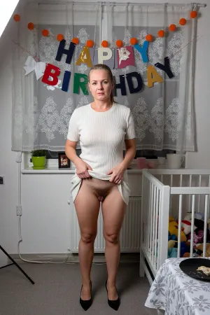 A candid, vertical photograph taken indoors with direct flash lighting, capturing a domestic birthday celebration scene. In the center stands a mature 50 years old woman with ponytail blonde hair and fair skin, wearing cream-colored, waffle-knit, short-sleeved knee-length dress paired with black high heels.She lifted the bottom edge of her dress with both hands up to her belly, showing that she was not wearing any panties and that her crotch was unshaven with natural, lush stubble. She faces the camera with a subtle, closed-mouth smile, Behind her, a large window is covered by sheer white curtains with a delicate floral lace pattern. A colorful banner hangs across the upper portion of the curtains, spelling out "HAPPY" in dark blue, gold, silver, red, and green letters, followed by a white bow cutout and "BIRTHDAY" in red, green, black, dark blue, and grey letters. An orange pom-pom garland is strung above the banner against the fabric. On the white window sill behind the woman, a green plant pot sits on the left, a small framed photograph in the center, and a pink cup and a white pot on the right. To the right of the woman, a white wooden crib or playpen stands against the wall, filled with various stuffed animals, with a white radiator visible behind it. The floor is covered in a neutral grey carpet. At the bottom right, the edge of a table is visible, covered with a white tablecloth featuring a grey floral pattern, holding a black plate with remnants of cake. The lighting is harsh and direct, characteristic of on-camera flash photography, casting a sharp, dark shadow of the woman and the banner against the curtains behind her. The image has a casual, snapshot aesthetic with slight digital grain and high contrast typical of low-light indoor photography.She lifted the bottom edge of her dress with both hands up to her belly, lush stubble