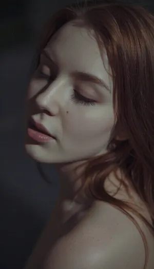 A muted, low-contrast sensual portrait in a very dimly lit, moody late night setting with all lights off. The scene is lit only by a very soft, underexposed, diffused ambient light source. The woman's sultry features are defined by subtle, soft-focus shadows. The image has a heavy film grain, and the colors are desaturated and washed out. Her red hair is soft and falls loosely over her delicate shoulders. Lidded eyes and soft full lips. The overall feel is intimate stillness and anticipation.