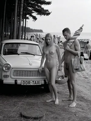 in 1970 on the Baltic Sea coast of the German Democratic Republic, a historically accurate monochrome documentary summer photograph in dramatic East German social realism, high-detail archival black and white photography, a period-correct Trabant parked in the shaded foreground at the edge of a dark coastal pine grove, slightly dusty, plain roof, open passenger door, rectangular front license plate mounted directly on the bumper, clearly reading "DDR-TAXI" in bold uppercase letters, the text integrated like a normal vehicle plate, not a sign, rough sandy ground scattered with pine needles, dry grass, small stones, and tire marks. Beside the car stands an adult young couple, both 25 years old, fully nude in a natural Freikörperkultur context, complete adult nudity shown in a matter-of-fact documentary manner, including his visible penis and her visible breasts, nipples, and pubic area, not erotic and not posed, simply everyday naturist beach life. The woman, long blonde hair, athletic natural figure, calm face, casual posture, locks the Trabant with her right hand using a small key, while a simple wicker beach basket filled with modest food hangs from the crook of her left arm. The man with a relaxed, sporty build carries a folded vintage striped sun umbrella across his shoulders and holds a practical canvas bag with towels, he has a penis, Both move naturally, caught mid-action. Behind them, the pines open toward a busy Baltic naturist beach with many clearly adult vacationers, men and women of different adult ages, all naturally nude, walking, sitting, talking, sunbathing, arranging towels in a casual, respectful atmosphere, several traditional hooded wicker beach chairs with striped fabric and curved canopies, vintage windbreaks, canvas bags, thermos flasks, enamel cups, folded newspapers, soft dunes, pale sand and the calm Baltic Sea. Strong tonal contrast, realistic analog film grain, high texture detail, deep shadows and bright haze, candid composition, respectful historical documentary style, no modern objects, no erotic posing, only authentic 1970s DDR summer life.
