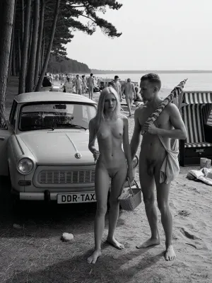 in 1970 on the Baltic Sea coast of the German Democratic Republic, a historically accurate monochrome documentary summer photograph in dramatic East German social realism, high-detail archival black and white photography, a period-correct Trabant parked in the shaded foreground at the edge of a dark coastal pine grove, slightly dusty, plain roof, open passenger door, rectangular front license plate mounted directly on the bumper, clearly reading "DDR-TAXI" in bold uppercase letters, the text integrated like a normal vehicle plate, not a sign, rough sandy ground scattered with pine needles, dry grass, small stones, and tire marks. Beside the car stands an adult young couple, both 25 years old, fully nude in a natural Freikörperkultur context, complete adult nudity shown in a matter-of-fact documentary manner, including his visible penis and her visible breasts, nipples, and pubic area, not erotic and not posed, simply everyday naturist beach life. The woman, long blonde hair, athletic natural figure, calm face, casual posture, locks the Trabant with her right hand using a small key, while a simple wicker beach basket filled with modest food hangs from the crook of her left arm. The man with a relaxed, sporty build carries a folded vintage striped sun umbrella across his shoulders and holds a practical canvas bag with towels. Both move naturally, caught mid-action. Behind them, the pines open toward a busy Baltic naturist beach with many clearly adult vacationers, men and women of different adult ages, all naturally nude, walking, sitting, talking, sunbathing, arranging towels in a casual, respectful atmosphere, several traditional hooded wicker beach chairs with striped fabric and curved canopies, vintage windbreaks, canvas bags, thermos flasks, enamel cups, folded newspapers, soft dunes, pale sand and the calm Baltic Sea. Strong tonal contrast, realistic analog film grain, high texture detail, deep shadows and bright haze, candid composition, respectful historical documentary style, no modern objects, no erotic posing, only authentic 1970s DDR summer life.