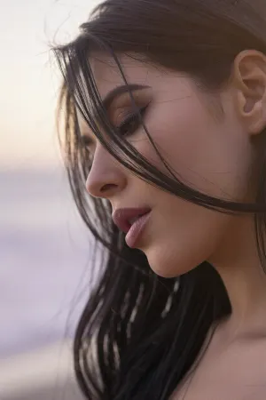 A tight intimate closeup portrait of a sultry Latina woman with an ethereal, soft-focus aesthetic, defined by diffused light. The Mexican woman's long elegant dark hair falls loosely across her face and obscures her sultry facial features while coyly hiding one eye behind the thick cascade of luxurious hair.
Long feathery eyelashes and dark smokey eye shadow with bold eyeliner.
Her plump luscious lips are a dark matte berry with soft pillowy creases. She has high sharp cheekbones.

The scene is enveloped in a powerful but diffused golden-hour glow from the side, which creates a luminous light bloom on her skin, dissolving sharp edges in an ethereal haze. The background is a very blurred, dreamlike pastoral pastel lavender-gray coastal sky, soft and bright. Soft focus on her luscious kissable lips.
The overall mood is an intimate serene sensuality. Shallow depth of field, subtle film grain, light leaks, vintage analog lens. This is a high-key photograph.
The subject is anchored to the right side of the image, using the rule-of-thirds compositional technique.