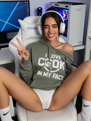 A casual "caught in the moment" photo of a 23 y.o.Moroccan amazigh tall very skinny woman with brown eyes sitting on a white gaming chair at a white standing desk with a curved monitor with blue desktop wallpaper, a white gaming PC with RGB, webcam glowing in the background. Cum is dripping from her forehead and nose and chin, the white gaming chair has a text "Team Suck" on the headrest. She is wearing an oversized olive green hoodie that hangs off one shoulder, She is wearing tight white pant shorts. She is wearing white gaming headphones with microphone. She is legs wide on the chair, wearing long white socks with Nike logo. On the hoodie there's a white big text "I LOVE COCK IN MY FACE", a logo with a penis inserted into a mouth. She is making a V sign, (peace sign), with the fingers of her right hand, eyes closed, big toothy grin showing her teeth, cute, playful expression