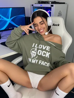 A casual "caught in the moment" photo of a 23 y.o.Moroccan amazigh tall very skinny woman with dark brown hair and brown eyes sitting on a white gaming chair at a white standing desk with a curved monitor with blue desktop wallpaper, a white gaming PC with RGB, webcam glowing in the background. Cum is dripping from her forehead and nose and chin and thighs, the white gaming chair has a text "Team Suck" on the headrest. She is wearing an oversized olive green hoodie that hangs off one shoulder, She is wearing tight white pant shorts. She is wearing white gaming headphones with microphone. She is legs wide on the chair, wearing long white socks with Nike logo. On the hoodie there's a white big text "I LOVE COCK IN MY FACE", a logo with a penis inserted into a mouth. She is making a V sign, (peace sign), with the fingers of her right hand, eyes closed, big toothy grin showing her teeth, cute, playful expression