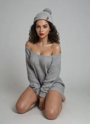 fullbody, Monochromatic studio portrait of a dark-skinned woman, late 20s/early 30s, kneeling on a pale gray background. She gazes towards the viewer, wearing a textured, off-the-shoulder chunky knit gray sweater and matching beanie with pom-pom. Large gold hoop earrings adorn her, with vibrant red lipstick on her lips. Dark curly hair peeks from the hat. Her hand gently touches her chin, creating a thoughtful, alluring expression. Soft, diffused lighting casts gentle shadows, suggesting intimacy, sensuality, and melancholic vulnerability. Classic composition, fashion/artistic photograph style, reminiscent of Peter Lindbergh and Herb Ritts, with a sophisticated, alluring mood.