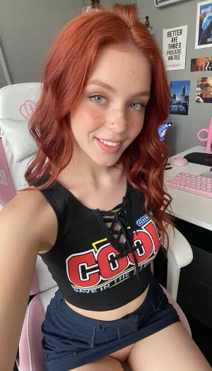 A young woman, 21yo, with striking red hair and freckles, seated in a gaming chair at a desk, is captured in a casual, selfie-style portrait, looking directly at the camera with a cheeky happy smile. Teeth visible.The composition is a close-up, centered on the woman, with the framing focusing on her upper body and face. The scene appears to be a personal bedroom, suggested by the gaming chair, desk, keyboard, and wall decorations. Elements are arranged to draw attention to the subject, with the background providing context without being distracting.The key subject is a young woman with abundant, wavy red hair that is loosely tied back. She has fair skin adorned with numerous freckles across her nose and cheeks. Her eyes are a striking blue-green, and her expression is calm and direct, meeting the viewer's gaze. She is wearing a black, sleeveless crop top with a lace-up front. The top features a bold graphic design with the word "COOL" in large red letters, accented with yellow and white. Below this, smaller text reads "SAVE IN THE FUTURE." She also wears a dark very short micro skirt, pulled up, panties visible. Hairy ginger pussyThe image is a photograph, likely taken with a smartphone in a selfie mode, given the perspective and framing. The technical execution is characteristic of amateur photography, with natural lighting and a focus on the subject. The style is contemporary and casual, reflecting personal aesthetics and potentially a connection to internet culture or gaming. The creative choice to include personal items like posters and gaming peripherals situates the subject within a recognizable modern environment.The setting is an indoor space, appearing to be a room with light grey walls. Visible elements include a white and pink gaming chair with a prominent logo, a white desk, a pink keyboard, and a computer mouse. The wall behind the woman is decorated with several framed posters, one of which has text reading "Better Things Ahead." Other posters depict urban landscapes and possibly anime-inspired artwork. The lighting appears to be a combination of ambient room light and possibly light from a screen, casting a soft, even illumination on the subject without harsh shadows, contributing to a relaxed and intimate mood.