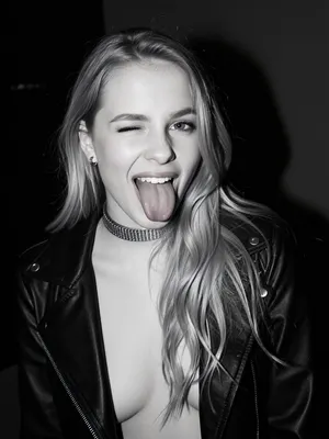 raw photo, A high-contrast black-and-white portrait captures a young woman in her twenties framed in a medium close-up shot from an eye-level perspective. She faces directly toward the camera with an animated, playful expression, winking one eye while sticking her tongue straight out. Her long, blonde hair is styled with soft waves and parted slightly off-center, flowing down over her shoulders. She has a slender build with a moderate chest visible beneath her attire. She wears an open black leather jacket over a low-cut dark top. A thick, metallic choker necklace sits at the base of her neck, accompanied by small hoop earrings visible near her jawline. The direct frontal flash creates sharp highlights on her face, collarbone, and the sheen of her leather jacket, casting deep shadows in the background. The background remains dark and largely uncluttered, drawing full focus to the subject. The monochromatic tonal range emphasizes bright highlights against rich, dark tones, giving the image a grainy, classic analog aesthetic reminiscent of candid flash photography.