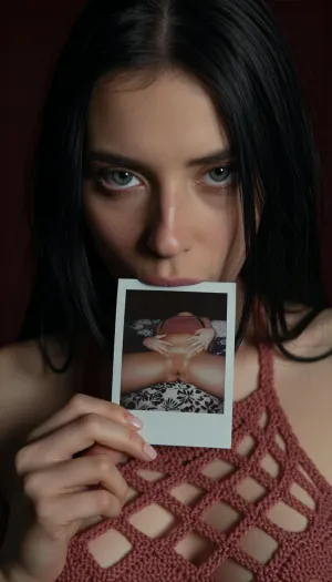 A close-up, high-quality of a young woman with straight long black hair, looking directly into the camera with an intense and slightly melancholic expression. Her light-colored eyes are fixed on the viewer. She is holding a small, square nude autoportrait instant polaroid photograph, (shows her lying on a patterned bedspread, hands positioned on her pubic area), between her lips. The lighting is moody and dramatic, with a focus on her face, creating a soft glow around her features while the background fades into deep shadow. She wears a burnt rose crocheted halter mesh top, (mesh top:1.9), (top grid:1.9), (transparent top:3), (open-knit texture:1.9), (((crocheted see through top:1.9))), (Her breasts in a top:1.5), (woman wears inesh top:2), (her the tits is covered by a wear:2), (breasts half covered:1.9), (tits covered:2), The style is reminiscent of stylized digital character art, capturing a cinematic and intimate moment with realistic textures on skin and hair, amateur aesthetic, high detail, sharp focus, atmospheric lighting.