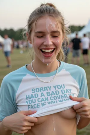 A candid, medium close-up photograph of a young tall skinny woman with fair skin and blonde hair pulled back in a messy ponytail, with loose strands framing her face. She is laughing heartily with her eyes closed and mouth open, displaying a joyful, genuine expression. She wears a white raglan t-shirt with light blue sleeves and a single-strand pearl necklace. The shirt features bold, light blue text reading "SORRY YOU HAD A BAD DAY YOU CAN COVER MY FACE IF YOU WANT." Two hands from the off are lifting the bottom hem of the shirt, groping her small breasts and twisting her nipples. a lot of cum dripping from her forehead and nose and lips and cheeks, The background is a soft-focus outdoor scene, likely a grassy field at a festival or park, with blurred figures of people standing in the distance. The lighting is natural daylight, creating soft shadows and highlighting her facial features. The photography style is realistic and documentary-like, with a shallow depth of field that keeps the subject sharp while blurring the busy background.