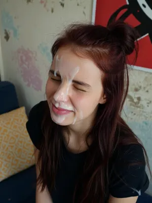 A young tall very skinny woman with long, dark reddish-brown hair, styled in a messy bun on top of her head with loose strands falling around her shoulders, her face covered in a white, viscous liquid. Her eyes are tightly squeezed shut, and her mouth is slightly open, revealing a hint of pink lips also covered in the liquid. She has a flushed complexion with some visible skin texture and small blemishes. She is wearing a dark, possibly black, t-shirt.

The background features a textured wall with subtle, faded pastel colors like light blue, pink, and beige, suggesting a worn or artistic finish. In the upper right corner, a portion of a vibrant red poster or artwork with abstract black patterns is visible. To the left, a yellow patterned pillow rests on a dark blue surface, possibly a bed or couch.

The lighting is direct and somewhat harsh, casting minimal shadows and highlighting the texture of the liquid on her face. The overall mood is intense and raw.

Captured with a close-up, slightly high-angle shot, using a wide aperture for a shallow depth of field, focusing sharply on her face. The image has a slightly grainy, candid, and unrefined aesthetic, reminiscent of early digital photography, with natural color grading.
