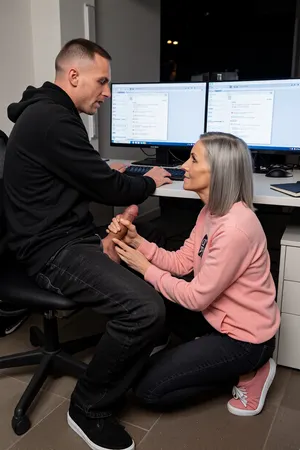 A man and a woman are in the middle of an open plan office. It is late night and the office is empty and dark. There is no one else. Around them is only darkness. The man is sitting on a chair in front of a Desk, working and looking at the screens. His Hands are on the keyboard on the table. On the desk is a Notebook and 2 illuminated flat screens with many open windows. The woman is squatting beside him. The man is 45 years old, tall and wears a black hoodie and black sneakers. His black jeans are wide open. His erected penis is visible. He wears a grey buzz cut, cropped to just 1mm. His head is nearly bald. He has a slender muscular body. His long and big and beautiful penis is hard erected. The woman is stroking his erected penis with one hand. Her other hand is grippinghis balls. The woman is 85 years old. She is clothed, she wears a pink Sweatshirt and black skinny jeans and pink sneakers. They are both fully visible. The man is looking to the screens with a smile.