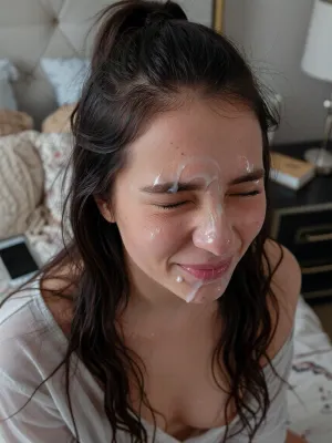A young woman with long, dark and loose brunette hair in a half bun, with loose strands falling around her shoulders, her face covered in a white, viscous liquid. Her eyes are tightly squeezed shut, and her mouth is slightly open, revealing a hint of pink lips also covered in the liquid. She has a flushed complexion with some visible skin texture and small blemishes. She is wearing a loose pajama top with visible cleavage.

The background features a bed in a lived in bedroom, with a very subtle bohemian style but a little bit hectic. There is a smartphone by her side and a side table with a book and side lamp.
The lighting is soft and cozy, casting minimal shadows and highlighting the texture of the liquid on her face. The overall mood is intense and raw.

Captured with a close-up, slightly high-angle shot, using a wide aperture for a shallow depth of field, focusing sharply on her face. The image has a slightly grainy, candid, and unrefined aesthetic, reminiscent of early digital photography, with natural color grading.