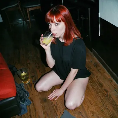 22 year old introvert redhead grunge girl with bangs sitting on the floor in a dark corner, drinking lemonade from a glass at a party in 1997, photo-realistic, compact camera, grainy