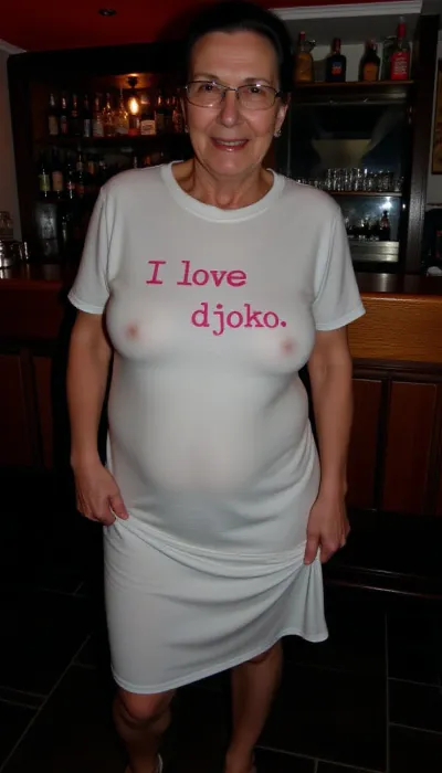 Candid amateur photograph, 55 year beautiful old smiling milf, (dark-haired ponytail), glassss, she wears a white t-shirt dress and low cut sandals, she is pulling her t-shirt dress up to her bulges belly revealing her shaved pubic hair, On her t-shirt dress is a Text printed in pink letters "I love djoko", she stands in front of a bar of a local pub in dim light, <lora:flux-age_sliders_latest_diffusers:.6>