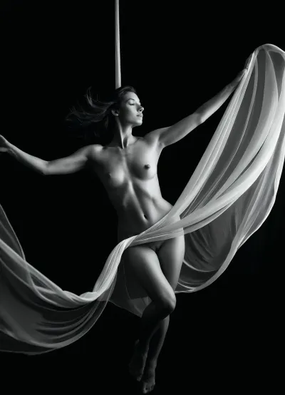 A black and white fine-art nude photography-style image of a woman suspended within flowing, sheer silk fabric that lifts and holds her in midair. The delicate, translucent material wraps around her body, contouring her natural form while creating