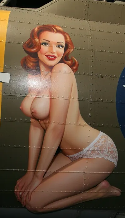 A vibrant and authentic vintage nose art painting stenciled onto the weathered olive-drab aluminum fuselage of a World War II B-17 bomber, captured in a slightly closer medium shot to emphasize the details. The artwork depicts a stunning 20-year-old redhead pin-up girl with a youthful face and exquisite features, painted in a lush oil style with bold brushstrokes. She is posed kneeling in a side profile view, completely topless, with her bare breasts visible from the side. She wears only delicate retro lace lingerie bottoms. She has a playful, knowing expression with classic red lips and perfectly styled retro wavy hair. The framing is focused from the waist up to just below her knees, bringing the viewer closer to the artwork while perfectly maintaining the kneeling profile pose. The painting is framed by metal rivets, panel lines, and weathered zinc-chromate scratches on the aircraft's metallic skin. Vibrant saturated colors, perfect vintage pin-up proportions, bold outlines suitable for military aircraft insignia, and high-contrast lighting.