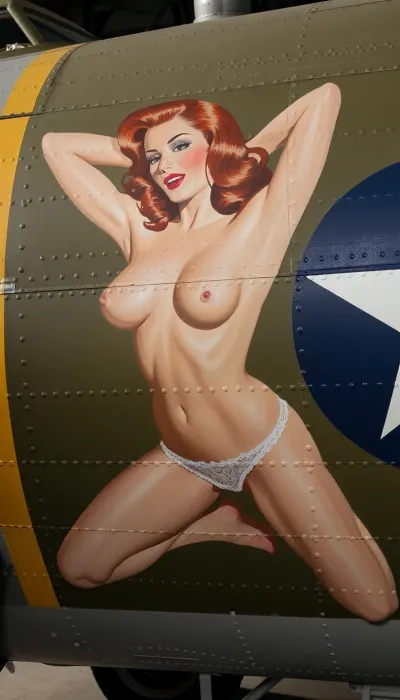 An authentic and vibrant vintage painting, stenciled onto the patinated olive-green aluminum fuselage of a World War II B-17 bomber, adorns the aircraft's nose. The artwork depicts a stunning 20-year-old redhead pin-up girl with a youthful face and exquisite features, painted in a lush oil style with bold brushstrokes. She poses kneeling in profile, her breasts completely bare, visible from the side. Her hands are behind her head. She wears only delicate retro lace lingerie panties. Her playful and mischievous expression, classic red lips, and perfectly styled retro waves make her a striking portrait. The painting is framed by metal rivets and panel lines. Vibrant, saturated colors, perfect proportions for a vintage pin-up, strong outlines reminiscent of a military aircraft insignia, and high-contrast lighting make the artwork stand out against the matte metallic background.