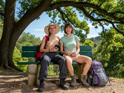 Two women sit on a green wooden bench in a sunlit forest clearing, framed by the lush green canopy of a large oak tree. The woman on the left wears a wide-brimmed beige sun hat, glasses, no top, braless small sized breasts, and dark hiking pants, with a red backpack visible behind her, she holds a smartphone and smiles warmly at the camera. The woman on the right, with short brown hair, wears a light green t-shirt, beige cargo shorts, and hiking boots, and holds a small notebook while smiling. Both are seated casually, exuding a relaxed, happy mood. The bench, with a green painted backrest, is mounted on stone supports. A purple and grey backpack rests on the ground beside the right woman. The background features dense foliage, a clear blue sky with light clouds, and a natural, rocky terrain. The lighting is bright and natural, suggesting midday sunlight filtering through the leaves, creating soft highlights and shadows. The image has a candid, documentary-style quality with a wide-angle perspective, capturing a genuine moment of outdoor leisure.