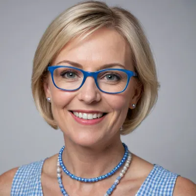 Close-up portrait of a smiling middle-aged woman with short blonde hair, wearing square glasses with blue frames, a light blue and white gingham dress, and a statement necklace of blue and white circular beads. The background is plain white. Natural lighting, head-and-shoulders shot, realistic style, high detail.