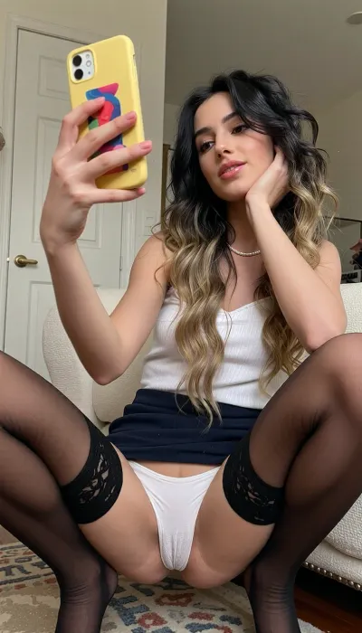 UHD, photorealistic. A young 6 feet tall very petite extremely skinny Argentinian woman with messy black wavy hair takes a selfie in a room, her gaze directed towards the phone screen as she poses in a squatting position.

The composition is a close-up, centered on the woman. She is framed from the mid-torso upwards, with her bent knees dominating the lower part of the image. legs spread, Her right arm is raised, holding a bright yellow phone with a colorful case, angled to capture her reflection. Her left hand rests on her chin, supporting her head in a pensive or playful pose. The framing emphasizes her facial expression and upper body, while her lower body and the surrounding environment provide context.

The key subject is a very skinny tall woman with fair skin, prominent cheekbones, and full lips. She has long, wavy blonde hair that cascades over her shoulders. Her eyes are looking towards the phone. She wears a white top and dark micro skirt, upskirt, skintight smooth white panties, complemented by tall, black, lace stockings. A delicate pearl necklace is visible around her neck. Her expression appears relaxed, with a slight pout to her lips and a happy content contemplative look in her eyes.

This image is a digital photograph, likely taken with a smartphone. The technical execution is that of a casual selfie, with a slightly soft focus that is characteristic of phone cameras, particularly when held at arm's length. The lighting appears to be natural, coming from an unseen source to the front, illuminating her face and the immediate surroundings. The style is informal and personal, capturing a moment of self-expression.

The setting is an indoor space, possibly a bedroom or living area. The background is slightly blurred but reveals details such as a white door with a brass handle on the left, and a textured, cream-colored chair or sofa. The floor has a rug with a subtle pattern. The lighting is soft and diffused, creating a warm and intimate atmosphere. The overall mood is casual and introspective, with a hint of playful confidence.