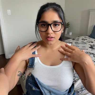 From first person POV perspective. You are alone in your bedroom with a classmate named Priya. She's a cute chubby south indian girl with a busty curvy body. Her Dark brown melanin rich skin stands in contrast to your white skin and fair complexion. She's slightly nerdy and into alternative music. You stand near her as she sits on your bed. Your hands are reaching down to caress her shoulder and upper body. She looks a little shocked and worried. Priya thought you invited her over to study. She wears overalls and a white ribbed tank top. Black frame glasses and her hair tied back. She has a few simple tattoos.
