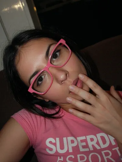 A low-angle, close-up shot of a woman with dark hair and PINK-rimmed glasses, looking directly at the camera with a neutral expression. She wears a pink t-shirt SUPERDRY SPORT and is positioned slightly off-center, with her head tilted. Her right hand is raised near her mouth, partially obscuring it. The lighting is dim and warm, creating soft shadows and a slightly grainy texture. The background is dark and indistinct, suggesting an indoor setting with minimal detail. The image has a candid, intimate feel, captured with a handheld camera, resulting in a slightly blurry and low-resolution quality. The composition emphasizes the subject's face and upper body, with a shallow depth of field that keeps her in focus while the surroundings remain out of focus. The overall mood is casual and personal, with a focus on the subject's direct gaze and subtle gesture.
