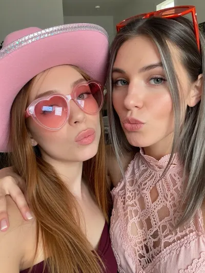 Create a square, close-up smartphone selfie of two fair-skinned people posing closely together indoors, a young woman on the left and an adult woman on the right. Their faces fill almost the entire frame, photographed at eye level with a slightly wide-angle front-facing phone camera. Both are making a playful “duck face” expression, with softly pursed lips, and are looking directly into the camera.

The young woman on the left has long, straight strawberry-blonde to light auburn hair, falling across her shoulders and chest. She wears an oversized pink cowgirl hat with a wide, gently curved brim. The hat has a pale blush-pink centre, vivid hot-pink edging and a subtle pearlescent or iridescent shimmer along parts of the brim. She also wears large, translucent pink heart-shaped sunglasses with rosy lenses and a shiny plastic frame. Her eyes remain faintly visible through the tinted lenses. A small bright white reflection from the phone or room lighting appears on the lower area of one lens. She is dressed in a dark plum or burgundy top, only partially visible at the bottom of the image.

The adult woman on the right has long, straight ash-blonde hair with darker roots, parted neatly through the centre and falling smoothly over both shoulders. She has light blue-grey eyes, softly shaped eyebrows, subtle neutral eye makeup, lightly contoured cheeks and glossy nude-pink lips. Include a tiny metallic nose stud on one nostril. Her expression is relaxed and playful, with her head tilted slightly toward the girl. She wears a dusty rose or muted mauve sleeveless lace top with a high scalloped neckline. The lace should have intricate floral and looping patterns, with slightly transparent sections revealing the fabric structure. A pair of narrow, bright coral-red or orange sunglasses rests on top of her head.

The woman has one arm wrapped naturally around the girl’s shoulders, drawing them close together. Her hand is visible near the far-right edge of the frame, resting comfortably behind the girl. Their cheeks are close, creating the feeling of an affectionate, spontaneous family selfie.

Use soft, warm indoor lighting with mild pink undertones. The light should be even across both faces, with gentle shadows and slightly smoothed skin similar to a subtle social-media beauty filter. Preserve realistic skin texture while maintaining the softly polished appearance of a phone selfie. Add faint catchlights in their eyes and glossy reflections on their lips and sunglasses.

The background is mostly out of focus and partially obscured by their heads. Include a pale grey wall or doorway behind them, a few dark shadowed areas, and a small glimpse of leafy indoor greenery in the upper-left corner. The upper-right background should be darker, with a bright coral-red horizontal object or reflection near the top edge. Keep the background visually quiet so the pink accessories and faces dominate the image.