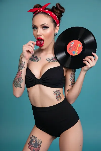 A studio portrait of a young woman in a classic pin-up style against a solid turquoise background, wearing a black retro swimsuit and a red bandana, covered in vibrant rockabilly-style tattoos. She is biting into a large cherry while holding a vintage vinyl record, with her eyes lined and bright red lips, she looks flirty and cheerful, and the vivid colors contrast with the solid-color background.
Studio photograph, a masterpiece, highest quality, pin-up brought to life
