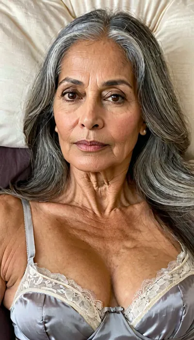 South Asian mature woman, 70 year old, full body view, (extreme oval shaped elongated face:1.3), ((waist long straight gray hair)), extremely messy hair, as woken up after a sleep, facial features The forehead shows fine to moderate horizontal wrinkles along with faint vertical lines between the eyebrows. Naturally full, gently arched eyebrows remain well defined but sit slightly lower due to mild age-related brow descent. The almond-shaped brown eyes remain expressive, though mild upper eyelid hooding, under-eye hollows, slight puffiness, and fine crow’s feet contribute to a mature appearance. straight, proportionate convex, smooth domed nose. The cheeks show some volume loss, creating a leaner mid-face with more noticeable nasolabial folds and some hollowing beneath the cheekbones. The lips are slightly thinner than in youth but remain well defined, fine vertical lip lines and shallow marionette lines extending from the corners of the mouth. The jawline appears softer with mild skin laxity along the lower face, neck displays visible platysmal bands, horizontal neck lines, and slight looseness beneath the chin. The skin retains a healthy, despite fine and moderate wrinkles around the forehead, eyes, and mouth. Long gray hair with natural gray at the temples and center part. Lying back in an oversized chair with her, legs slightly open, black nylon panties, and a lose white lace night dress, full body view.
