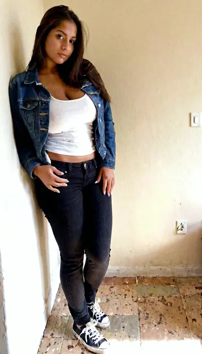 Ultra-realistic smartphone photograph of a 25-year-old adult Hispanic woman with a naturally slim-curvy feminine figure, warm olive skin, long dark brown hair and expressive brown eyes. She is wearing a denim jacket over a white top with black trousers and casual sneakers. She is leaning casually against a wall in a minimalist living room, looking directly at the camera with a relaxed expression. Natural indoor daylight, handheld smartphone camera, realistic exposure, authentic skin texture, detailed denim fabric, natural shadows, true-to-life colors, candid photography, highly photorealistic.