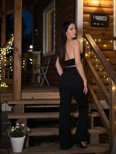 A beautiful slim young woman with long dark hair, wearing a sleek black sleeveless crop top and matching high-waisted black trousers, standing on a rustic wooden staircase. The scene is set at night against the exterior of a cozy wooden cabin with horizontal timber planks. Warm golden fairy lights are draped elegantly along the wooden handrail and stairs, creating a soft, magical glow and a cozy atmosphere. She is captured in a three-quarter profile, looking towards the camera with a gentle expression. In the foreground, a small white pot with delicate flowers sits on a step. The lighting is cinematic and warm, with a soft bokeh effect in the background, high detail, realistic textures, 8k resolution, atmospheric night photography.