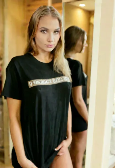 exact woman from the reference photo, identical face, standing in front of a large mirror, looking at her own reflection, oversized dark t-shirt, bedroom, soft lighting.
