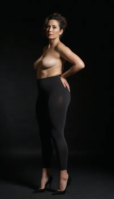 A full-length high-fashion editorial photograph of a poised 40-year-old woman with a dramatic, exaggerated pear-shaped silhouette. She has a slender, flat saggy chest that contrasts sharply with wide, opulent, curvaceous hips and a full, rounded posterior. She is dressed in understated, casual everyday attire—a simple fitted cotton tee and tight-fitting leggings—that clings to her form. She stands poised in black, needle-thin stiletto high heels that dramatically arch her feet. The lighting is cinematic rim lighting, carving her lower body's silhouette against a deep, midnight-black velvet backdrop. Hyper-realistic skin textures, visible pores, natural skin folds, and subtle cellulite on the thighs for raw authenticity. 8k resolution, shot on Phase One XFM, sharp focus on the hip-to-waist contrast, soft-focus background, sultry and sophisticated mood