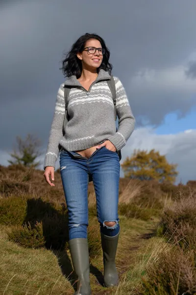 A low-angle, full-body shot of a smiling woman hiking up a grassy hill covered in heather. She has shoulder-length dark wavy hair and wears rectangular glasses. Her expression is joyful and relaxed, looking slightly off-camera. She wears a grey and white patterned quarter-zip knit sweater, fitted blue jeans with a small tear at the knee, and olive green rubber boots. Her jeans a pushed down to her knees, revealing her hairy vagina, so that her labia are presented to the viewer. A backpack strap is visible over her shoulder, one hand rests casually in her pocket. The setting is an autumnal moorland with sparse trees showing yellow foliage under a dramatic, cloudy sky with patches of blue. Lighting is natural daylight, soft but directional, highlighting her face and sweater texture. The composition emphasizes her upward movement against the expansive sky. Style, realistic outdoor photography, candid moment, shallow depth of field blurring distant trees, sharp focus on subject, earthy color palette with muted greens, greys, and blues. Capture the essence of casual adventure and connection with nature.
