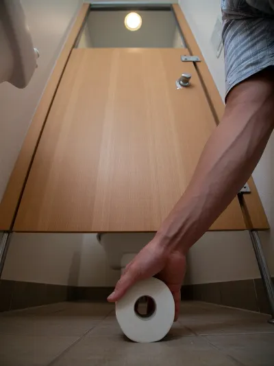 A dramatic low-angle worm's-eye view from the floor inside a narrow toilet stall, looking upward. A human hand holding a roll of toilet paper is extending inside through the wide bottom gap underneath the wooden stall door. The camera rests on the tiled floor, capturing the wooden door panel, the hand in motion, and the bright recessed ceiling light glowing directly overhead in the background. Cinematic vertical perspective, realistic shadows.